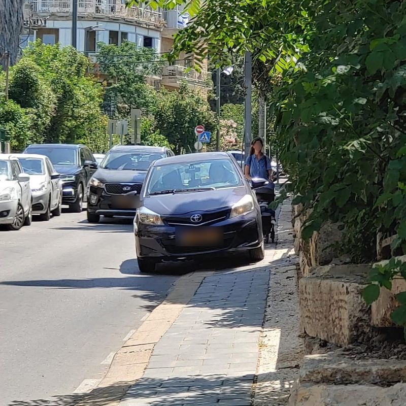 רכב עולה על מדרכה צרה, ואישה עם עגלת תינוק נדחקת לצד